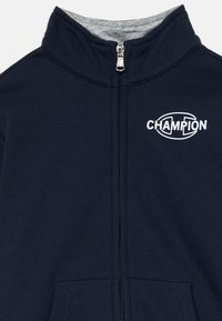 Chaqueta navy azul con cremallera y cuello alzado, bolsillos frontales y el logo blanco de Champion en el pecho superior izquierdo.