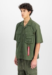 Camicetta a maniche corte verde oliva in cotone, con due tasche sul petto, un'etichetta con scritte rosse e dettagli ricamati sulle spalle.