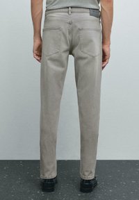 Jeans dritti grigi chiari con una texture opaca, due tasche posteriori e una patch in pelle nera sulla vita, abbinati a scarpe nere.