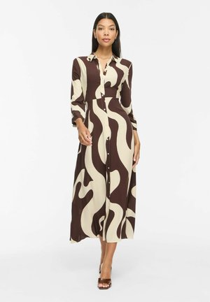 Femme portant une robe à manches longues boutonnée avec des motifs abstraits crème et marron, debout devant un fond blanc uni.