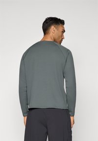 Chandail à manches longues en rib sweat dans une teinte vert foncé, avec un col rond et un tissu texturé. Modèle vu de dos, associé à un pantalon noir.