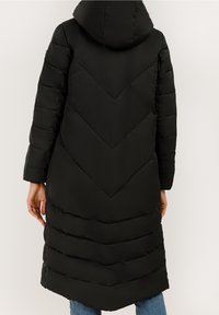 Abrigo negro acolchado con capucha, con un patrón de tejido en forma de chevrón, mangas largas y diseño hasta la rodilla. Textura suave y brillante.