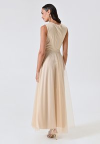 Beige Abendkleid mit taillierter Oberlage, fließendem Rock und transparenter, glitzernder Überlagerung. Der Rücken hat eine glatte Verarbeitung und dezente Nahtdetails.