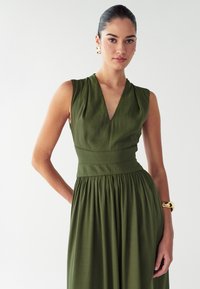 Vestido verde sin mangas con escote en V, corsé ajustado y falda plisada. Tejido suave con una textura sutil. Pulsera de brazalete dorado.