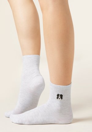 Beine tragen weiße gerippte Knöchelsocken, auf einer Socke ist nahe dem Knöchel ein kleines schwarz-weißes Hundegesicht eingestickt.