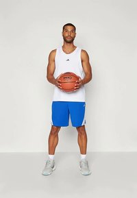 Valge sportlik särk Adidas'i logoga, sinised lühikesed püksid valgete triipudega, hoiab korvpalli, jalas hallid spordijalatsid. Siseruum.