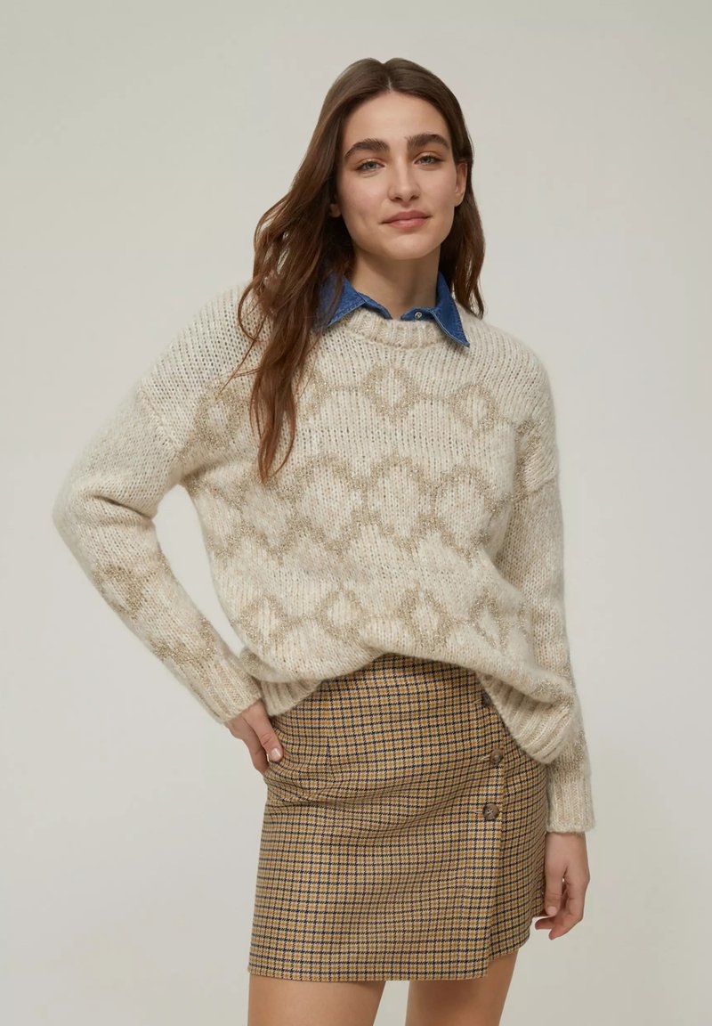 Maglione a maglia crema con motivo testurizzato, indossato sopra una camicia blu con colletto. Il modello indossa una gonna a quadri marrone con dettagli a bottoni.