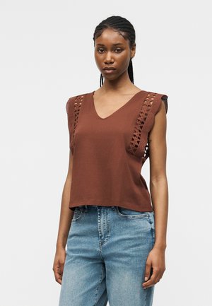 Kvinde med fletninger iført brun ærmeløs top med blonderdetaljer på skuldrene og lyseblå højtaljede jeans, stående mod en ensfarvet baggrund.