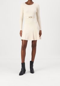 Robe crème côtelée à manches longues, corsage ajusté, jupe évasée et ceinture dorée en accent. Associée à des bottines noires à talons.