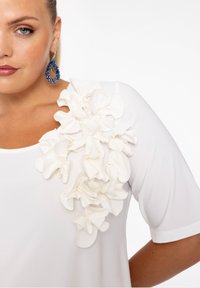 Blouse blanche avec des fleurs en tissu tridimensionnel sur une épaule, texture semi-transparente et manches courtes. Le modèle porte des boucles d'oreilles bleu drop.