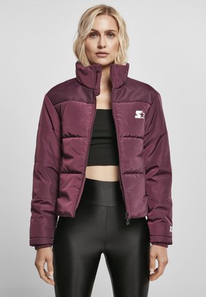 Blonde Frau trägt eine gestepptes Pflaumen-Jacke mit Reißverschluss über einem schwarzen Crop-Top und schwarzen glänzenden Leggings, steht vor neutralem Hintergrund.