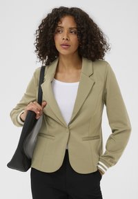 Olive green blazer in a soft fabric with a tailored fit. Features two front pockets and white-striped cuffs. Worn over a fitted white top.