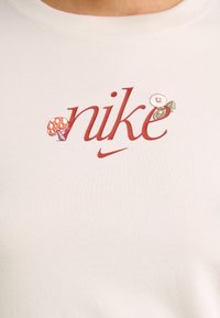 Crème kleurig t-shirt met geborduurd "Nike" logo in het rood, aangevuld met rode en witte paddestoel- en bloemaccenten.