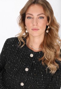 Chaqueta negra texturizada con hilo plateado, acentos de botones de perlas, collar de perlas en capas y aretes de aro dorados con un gran detalle de perla.