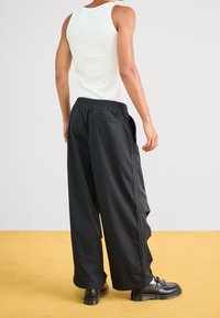 Schwarze, locker sitzende Jogginghose mit elastischem Bund und Seitentaschen, kombiniert mit einem weißen Tanktop und schwarzen Schuhen.