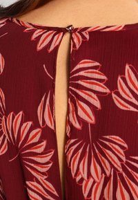 Robe bordeaux avec un motif floral rouge et rose. Présente une ouverture en forme de goutte d'eau dans le dos, maintenue par un petit bouton en métal. Tissu texturé.