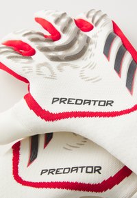 Gants de gardien de but blancs et rouges avec une texture antidérapante sur les doigts, un design en mesh, des rayures sur le côté et une marque "PREDATOR" sur le poignet.