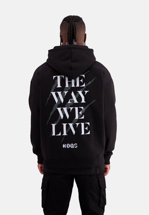 Schwarzer Hoodie mit einem großen Grafiktext "DIE ART, WIE WIR LEBEN" auf der Rückseite, versehen mit Kratzern. Weicher Stoff, entspannter Schnitt.