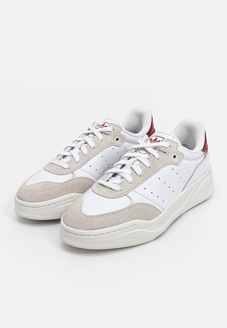 Zapatillas bajas en blanco y beige con cordones, lados perforados, lengüetas en color burdeos en el talón y el logo de Adidas en la lengua y la suela, lado a lado sobre un fondo blanco.
