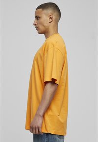 T-shirt en coton orange à coupe décontractée, à manches courtes et à encolure ronde, porté avec un jean en denim bleu. Vue de profil.
