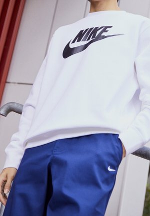 Osoba w białej bluzie Nike i niebieskich spodniach Nike stoi przy metalowej poręczy na tle ściany budynku.
