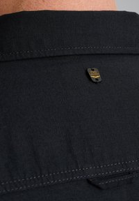 Cuello de camisa negro con un acabado de tejido texturizado, que presenta una etiqueta metálica con el texto "PME LEGEND" adjunta cerca de la parte posterior.