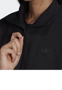 Black zip-up sweatshirt made of soft fabric; features a high collar and subtle logo on the chest. Zipper pull held by a hand.