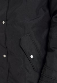 Zwart stoffen jack in close-up met een klepzak en een zilveren drukknoop, en een deel van een mouw met stikdetails.