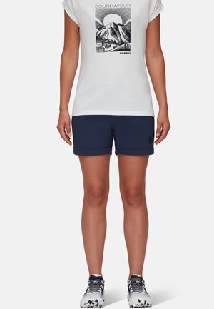 Kvinde iført hvid t-shirt med bjergmotiv og sorte shorts, stående med armene afslappet langs siderne mod en ensfarvet baggrund.