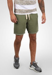 Shorts en coton verts avec des ourlets roulés, dotés d'une taille à cordon. Portés avec des baskets grises et une chemise à rayures blanches et dorées.