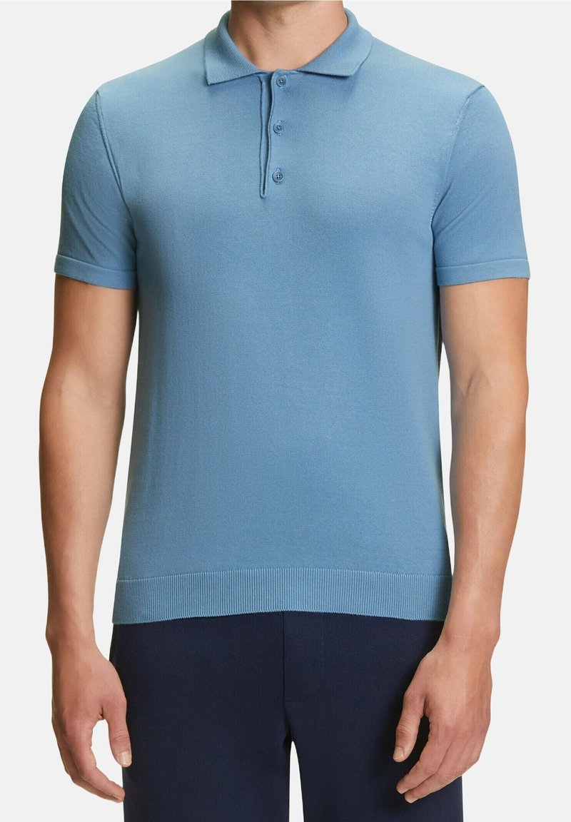Homme portant un polo à manches courtes bleu clair avec une patte de boutonnage à trois boutons et un pantalon foncé sur fond blanc.