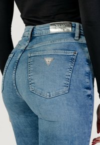 Jeans in denim azzurro chiaro con vita alta e tasche posteriori. Un logo argentato "GUESS" si trova in corrispondenza della vita e sulla tasca destra.