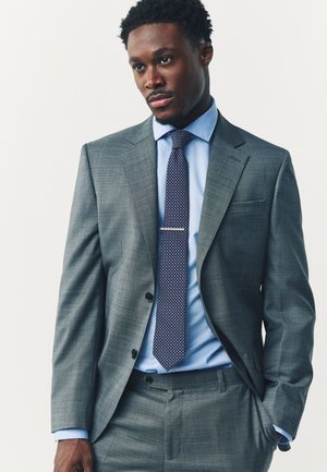 Mann in grauem Anzug mit blauem Hemd und gemusterter Krawatte, der mit einer Hand in der Tasche vor einem einfarbigen Hintergrund steht.