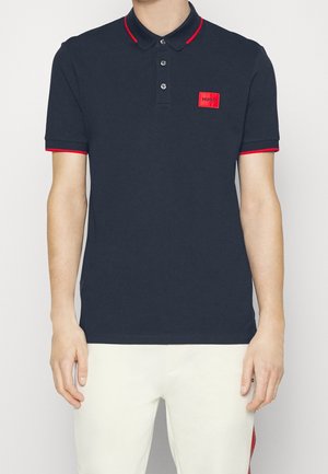 Homme portant un polo bleu marine avec des bordures rouges sur le col et les manches, patch logo Hugo rouge sur la poitrine, assorti à un pantalon blanc.