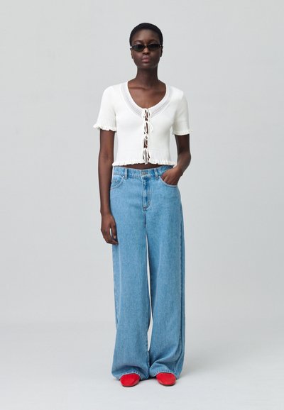 Top corto in maglia bianca con maniche corte a volant e dettaglio con fiocco. Jeans a vita alta azzurri larghi. Scarpe rosse completano l'outfit.