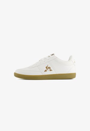 White sneaker with a smooth leather upper, perforated details, and a tan rubber sole. Features a gold logo on the side and matching accents.