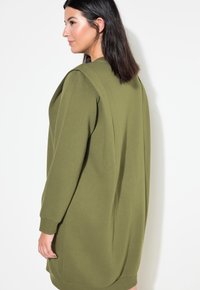 Sudadera oversize de color verde oliva, fabricada en un tejido suave, con mangas largas y un corte relajado. Presenta un detalle de hombro plegado y puños acanalados.