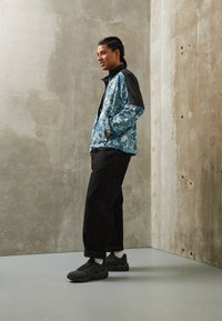 Veste imprimée florale bleue colorée avec des accents noirs, pantalon cargo noir et chaussures noires. Texture lisse, coupe décontractée, en position debout.
