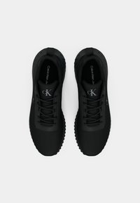 Black athletic shoes with a textured upper, round toe, flat laces, and patterned soles. Features branding on the tongue and side.
