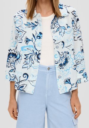 Vrouw die een wit topje draagt, een lichtblauw bloesje met bloemenprint en driekwart mouwen, en lichtblauwe broek met hoge taille en voorzak.