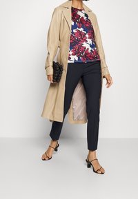 Béžový trenčkot přes květinový top v námořnické modři, červené a bílé barvě, spárovaný s tmavými kalhotami a černými sandály na vysokém podpatku. Černá kabelka.