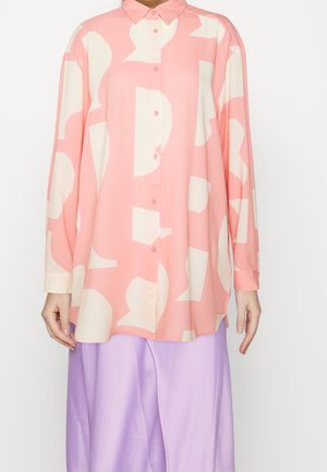 Vrouw draagt een loszittend roze overhemd met abstracte witte vormen, gecombineerd met wijde lila broek.