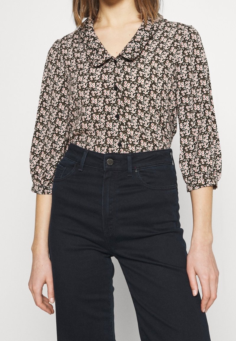 Blusa nera con stampa floreale rosa, colletto tondo e maniche lunghe a sbuffo. Abbinata a jeans blu scuro a vita alta.