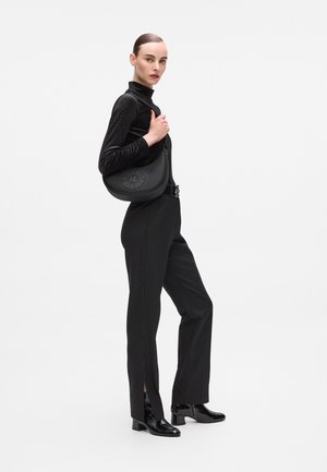 Black shoulder bag with a smooth texture and rounded shape, featuring a logo in studded design. Model wears a black, fitted turtleneck and pants.