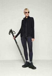 Donna che indossa abbigliamento sportivo nero e occhiali da sole, con in mano due bastoni da trekking neri, in piedi su uno sfondo chiaro e semplice.