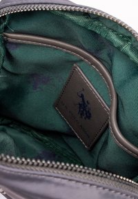Szary materiałowy worek z zamkiem błyskawicznym. Wnętrze ma ciemnozieloną, pasiastą podszewkę oraz skórzaną metkę z napisem "U.S. POLO ASSN." w ciemnoniebieskim kolorze.