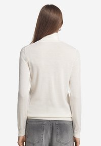 Cremfarbener, leichter Rollkragensweater mit langen Ärmeln. Hat ein nahtloses Design und gerippte Bündchen. Kombiniert mit grauen Jeans.