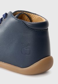 Detail obuvi pro miminka v námořnické modré kůži s hnědou podrážkou a vyraženým jablečným logem na patě.