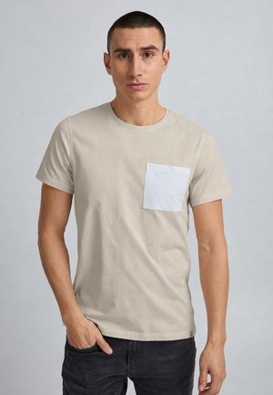 Mand med kort mørkt hår iført en beige t-shirt med en lyseblå brystlomme og sorte bukser mod en ensfarvet baggrund.