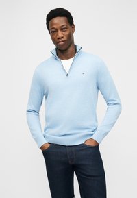 Άνδρας που φορά ανοιχτό μπλε πουλόβερ με φερμουάρ πάνω από λευκό πουκάμισο και σκούρο μπλε τζιν, στέκεται με τα χέρια στις τσέπες μπροστά σε απλό φόντο.